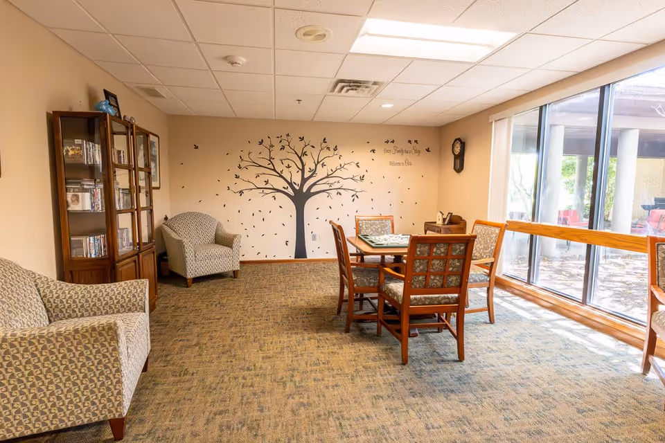 A cozy common area in an assisted living facility with patterned armchairs, a wooden table with four chairs, a glass-front cabinet filled with books and DVDs, and a large window letting in natural light. The wall features a decorative tree decal with birds and the phrase 'Every Grandday is a Story Welcome to Elison'.