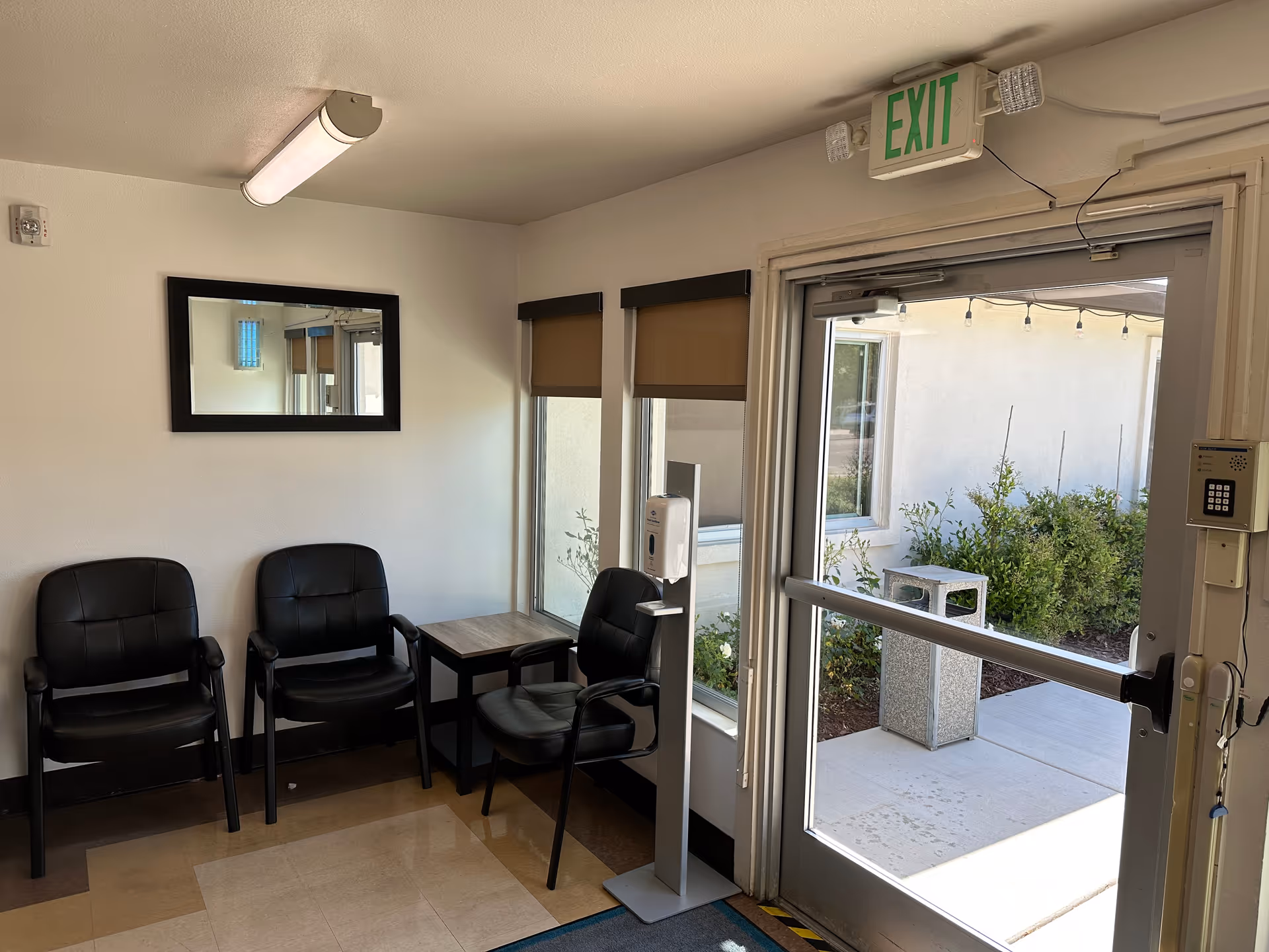 Small waiting area by an exit door with three black chairs, a side table, a mirror, and a hand sanitizer stand.