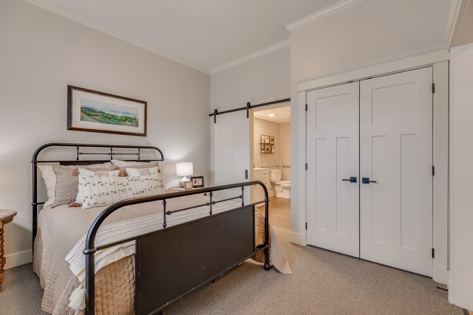 A cozy bedroom with a metal frame bed adorned with multiple pillows and a beige quilt. A small wooden side table with a lamp and picture frame is next to the bed. A framed landscape painting hangs above the bed. To the right, there is a white double-door closet and an open sliding barn door leading to a bathroom with a visible toilet and vanity.