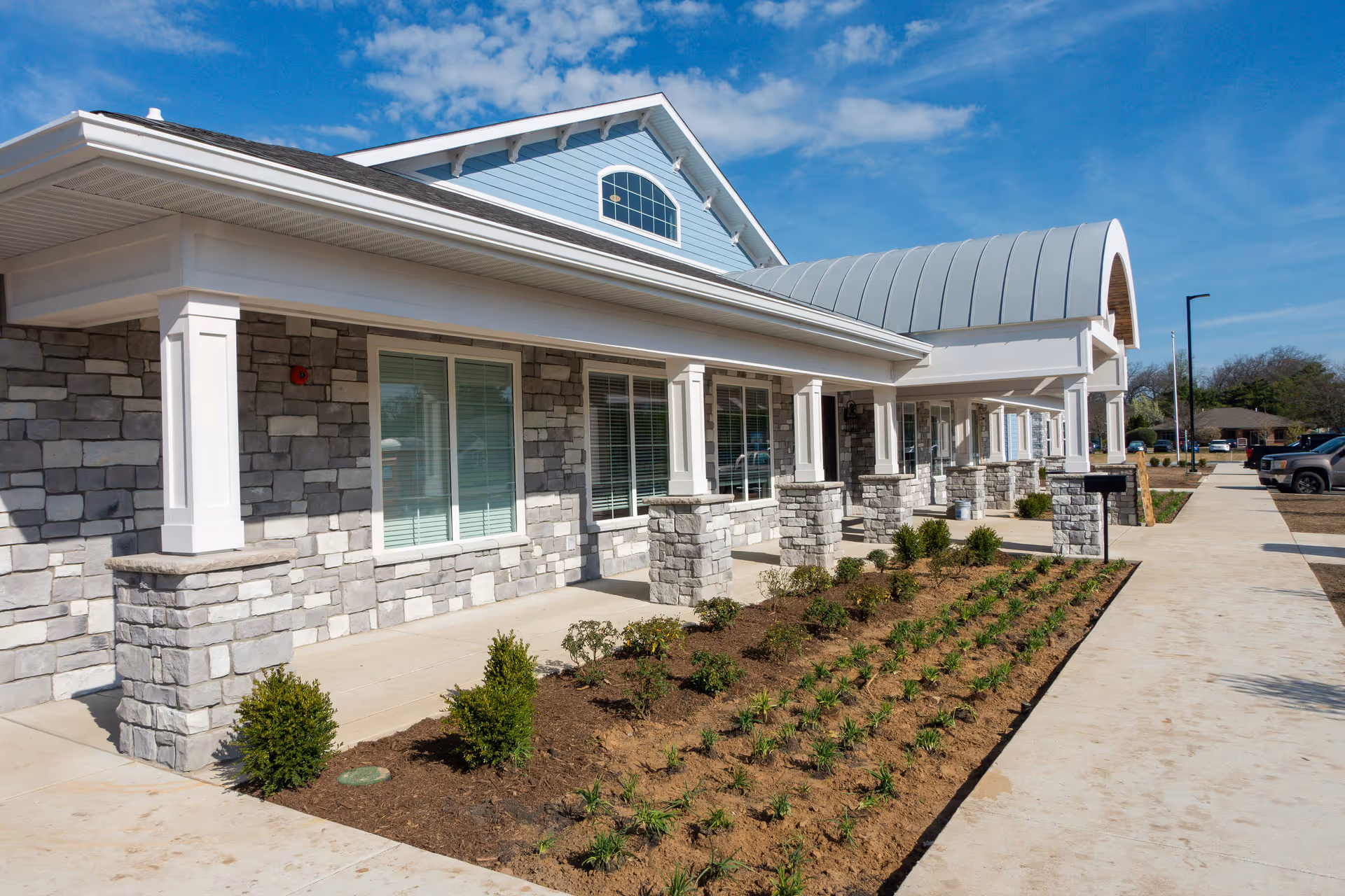 Front exterior of a stone-and-siding senior living building with a covered walkway, columns, windows, and newly planted landscaping along a sidewalk.