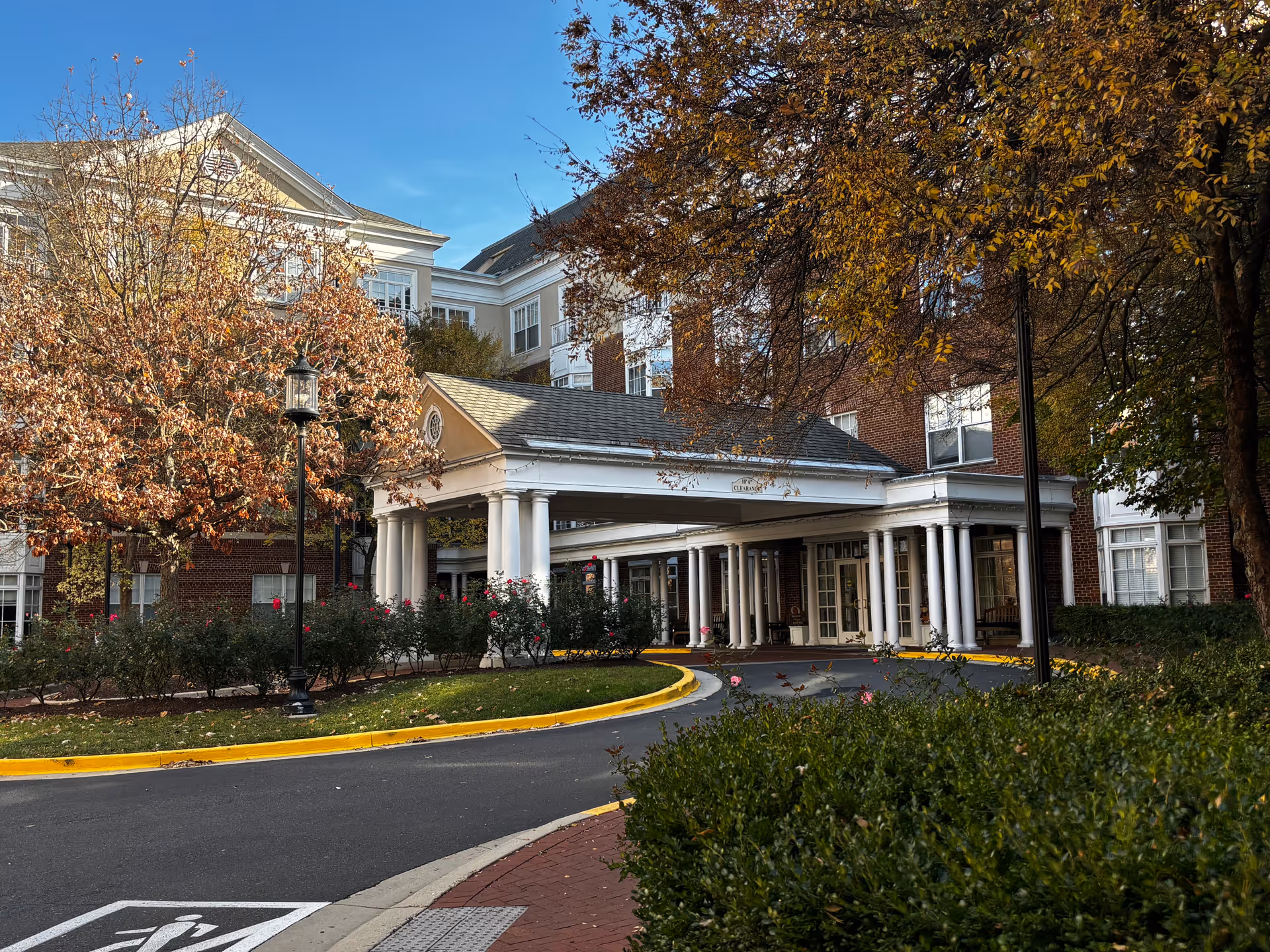 Front entrance of an assisted living community with a covered portico and columns, a circular driveway, and trees with autumn foliage.