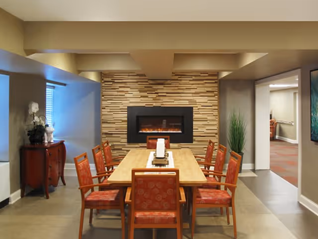 A dining room with a rectangular wooden table surrounded by eight red cushioned chairs. The back wall features a modern electric fireplace set into a textured wooden panel. To the left, there is a small red cabinet with decorative items on top, and to the right, a tall green plant. The room has a neutral color palette with beige walls and ceiling.