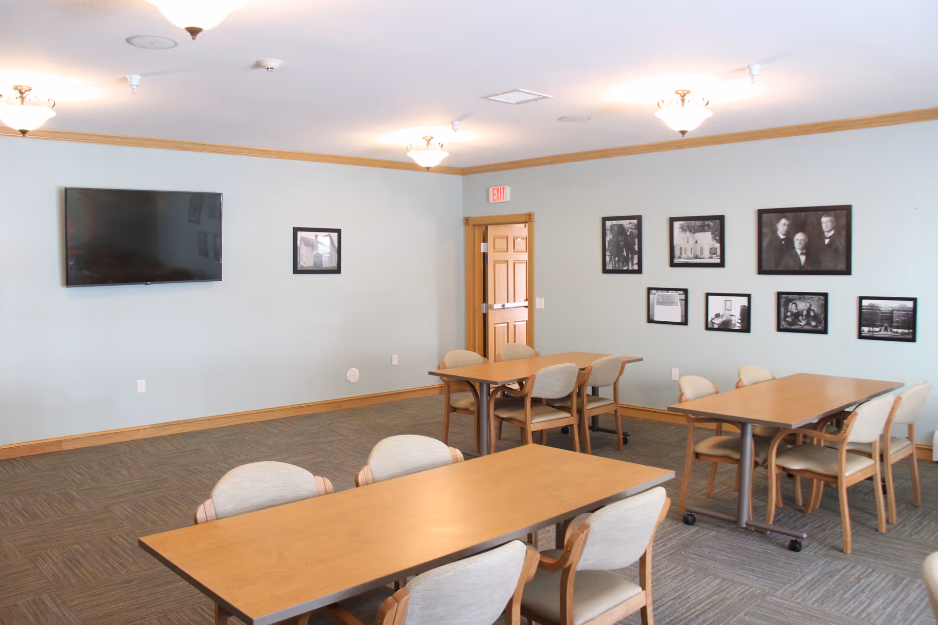 Large common dining/activity room with several wooden tables and upholstered chairs, a wall-mounted TV, framed photos, and ceiling lights.