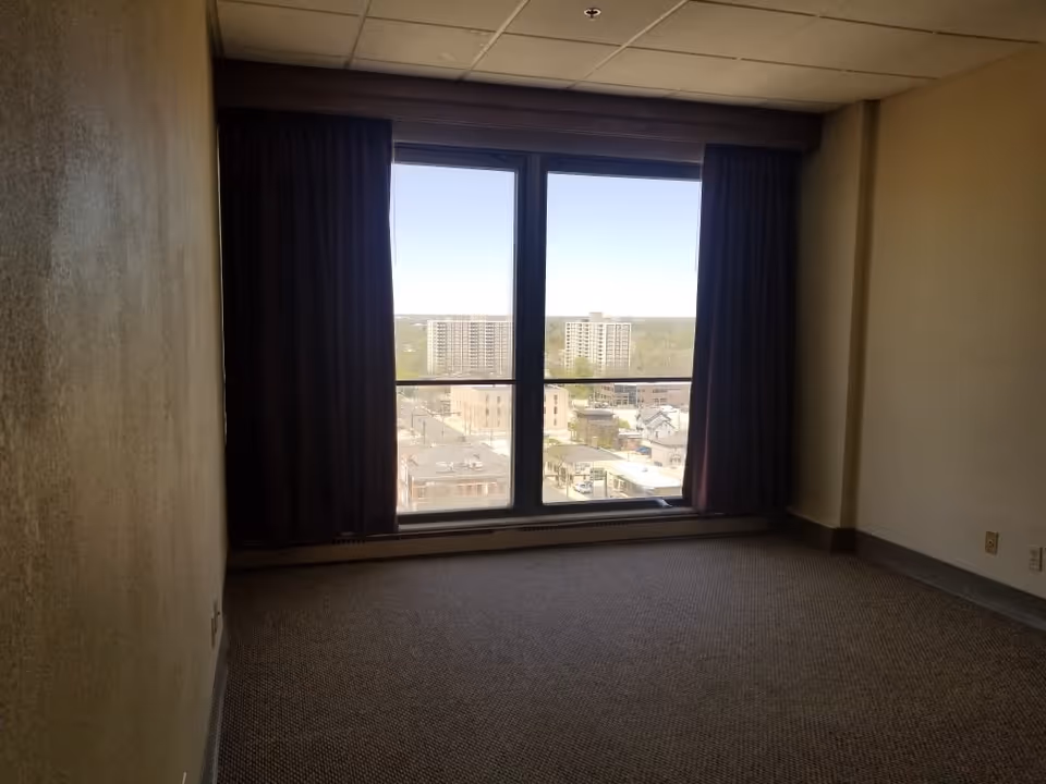 Empty room with beige walls and carpeted floor featuring a large window with dark curtains partially drawn, showing a cityscape with buildings and trees outside.