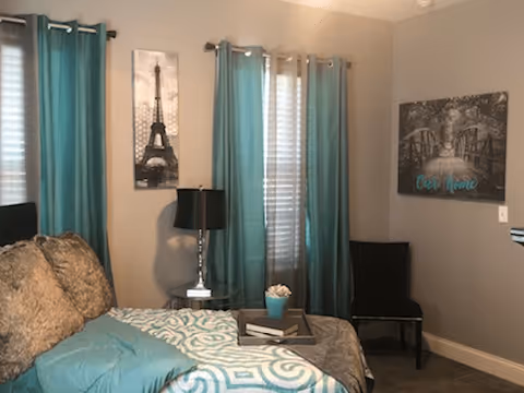 A cozy bedroom with a bed covered in teal and beige bedding, a small side table with a lamp and decorative items, teal curtains on the windows, and wall art including an Eiffel Tower print and a black and white photo with the words 'Our Home'.