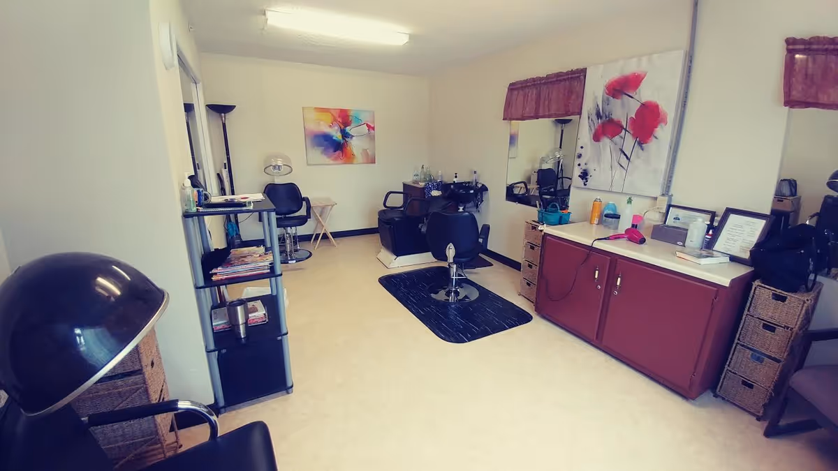 Interior view of a small hair salon or beauty room with salon chairs, a hair dryer, a mirror, a cabinet with hair products, and colorful artwork on the walls.