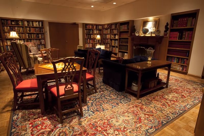 A cozy library room with wooden bookshelves filled with books lining the walls, a wooden table with four chairs featuring red cushions, a patterned area rug, two armchairs, a dark blue sofa, and a wooden side table with a lamp and decorative items. The room is warmly lit with lamps and recessed ceiling lights.