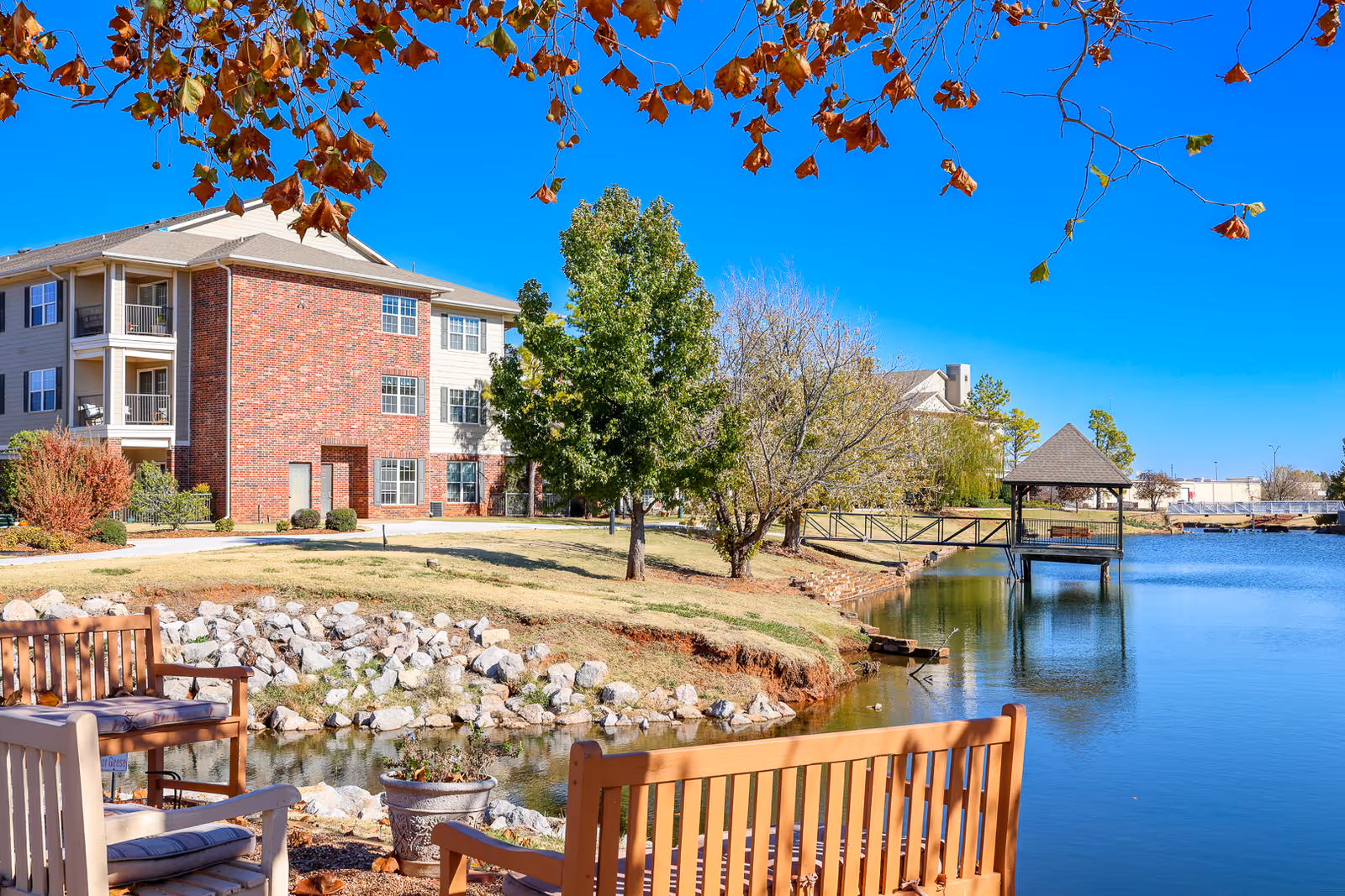 Outdoor view of Elison Independent Living of Statesman Club featuring a calm pond with a wooden gazebo on a dock, surrounded by trees and a three-story residential building with brick and siding exterior. Wooden benches and chairs are placed near the water's edge under a tree with autumn leaves.