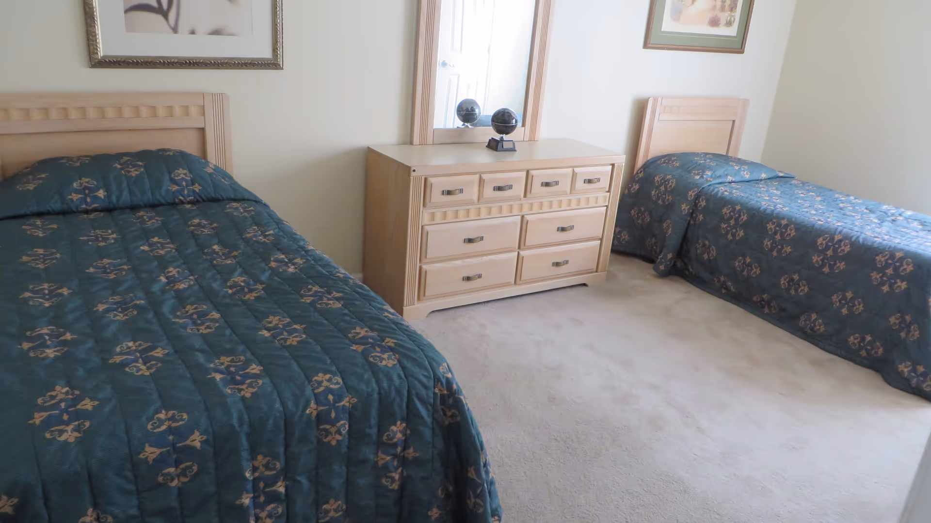 A bedroom with two single beds, each covered with a dark green patterned bedspread. Between the beds is a light wood dresser with multiple drawers and a mirror on top. The room has light-colored walls and carpet, with framed artwork hanging above each bed.