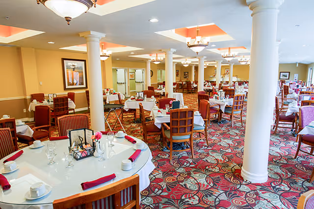 Spacious dining room with multiple round and square tables set with white tablecloths, red napkins, cups, and glasses. The room features patterned carpet, white columns, warm yellow walls, and ceiling lights with chandeliers.