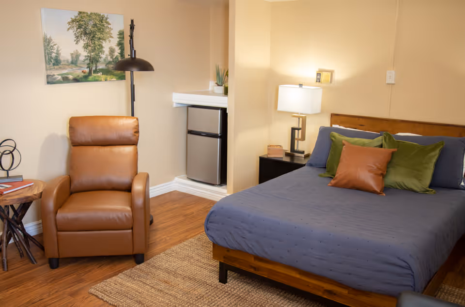 A cozy bedroom featuring a bed with blue bedding and accent pillows, a brown leather recliner, bedside table with lamp, a small refrigerator in the corner, and a landscape painting on the wall.