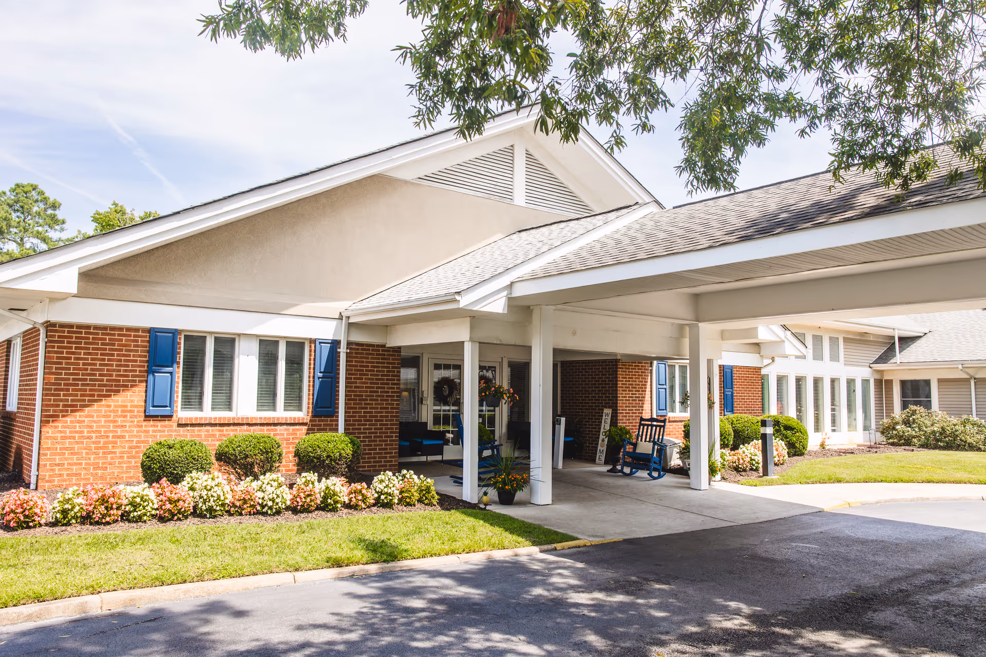 Exterior view of Churchland Nursing Home showing a brick building with blue window shutters, a covered entrance with white pillars, rocking chairs, and well-maintained landscaping including bushes and flowers under a partly cloudy sky.