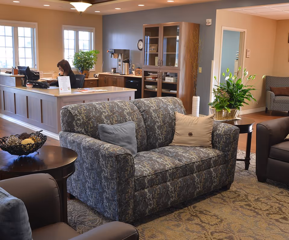 A cozy senior living facility common area with a patterned loveseat and armchairs arranged around a coffee table. In the background, a woman is seated at a reception desk with a computer and phone. The room features warm lighting, a cabinet with glass doors, potted plants, and large windows letting in natural light.