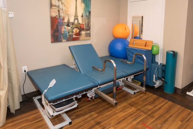A therapy room with a blue adjustable treatment table, exercise balls in orange and blue, a mirror on the wall, and a colorful painting of the Eiffel Tower. The room has wood flooring and beige walls.