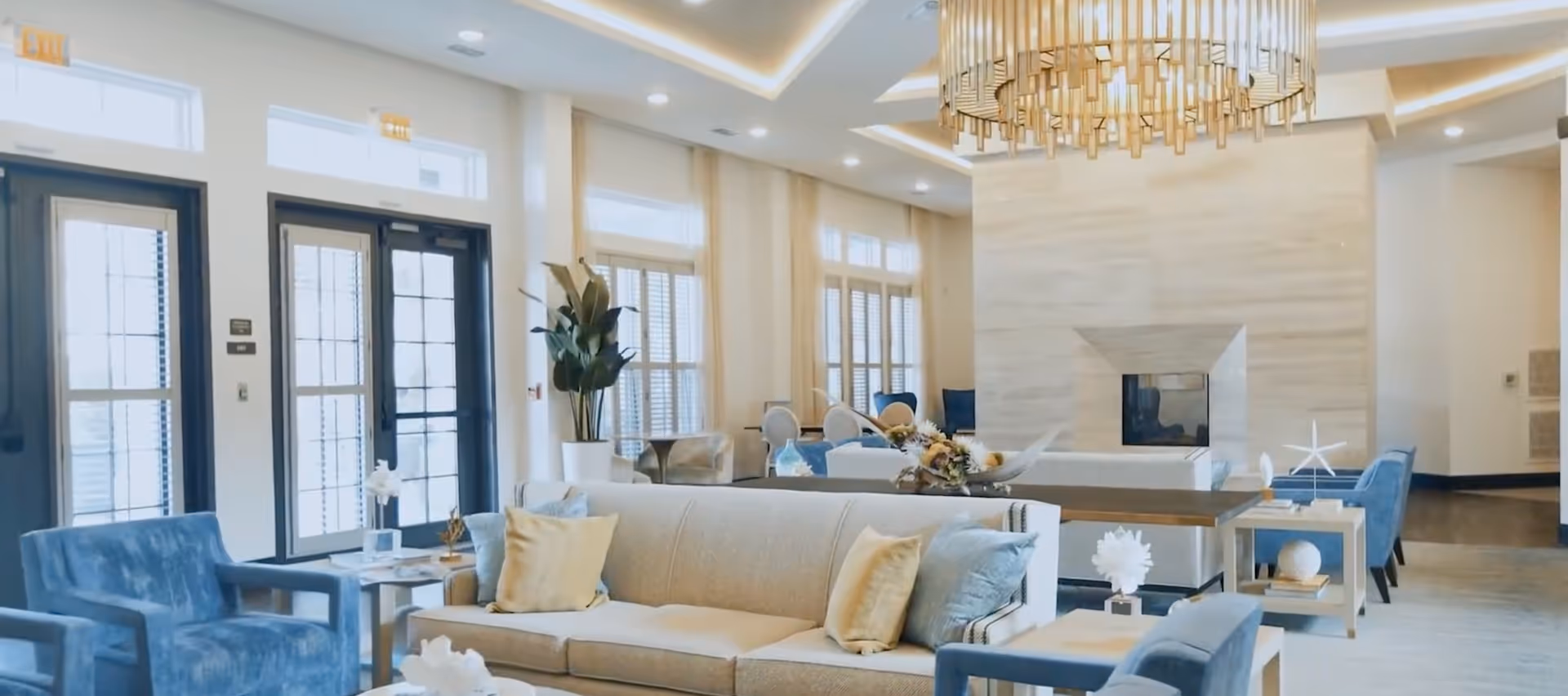 Bright upscale lounge with sofas and armchairs centered around a fireplace, a decorative chandelier, and large windows/doors.