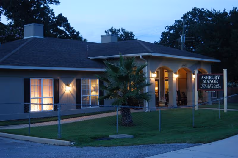 Exterior view of Ashbury Manor, a single-story assisted living facility, during dusk. The building has a dark roof, beige walls, and several lit windows. There is a small palm tree and a chain-link fence in front of the building, along with a sign that reads 'Ashbury Manor Specialty Care Assisted Living.'