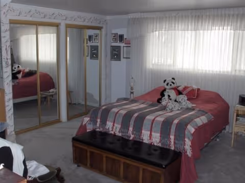 Bedroom with a bed covered in a red and gray blanket topped with stuffed panda toys, mirrored closet doors, a window with vertical blinds, and a bench at the foot of the bed.