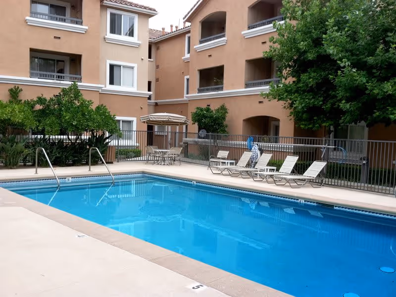 Outdoor swimming pool with lounge chairs and a fenced patio beside a multi-story beige apartment building.