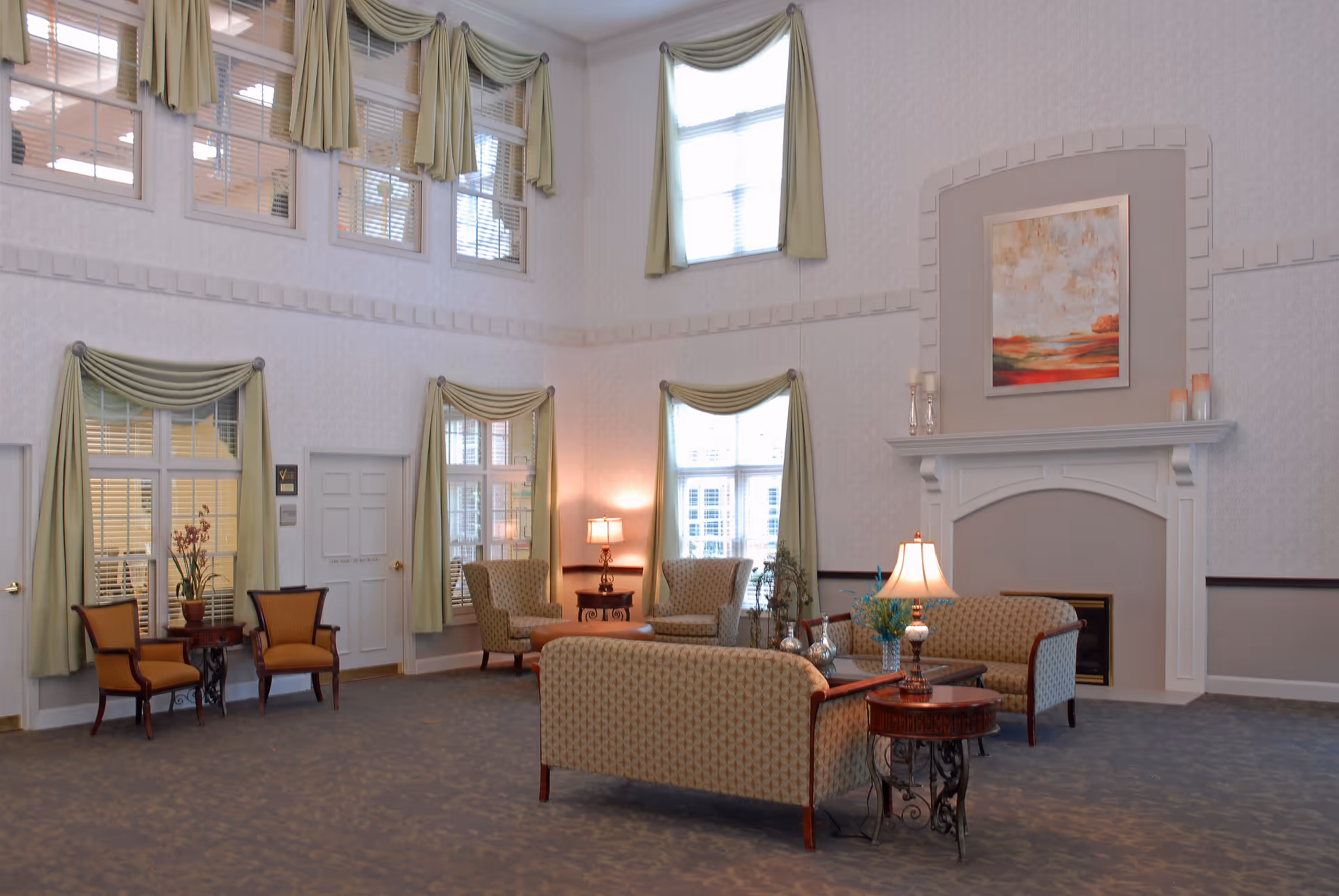 A spacious senior living facility common area with high ceilings and large windows adorned with green curtains. The room features comfortable patterned sofas and armchairs arranged around a fireplace with a painting above it. There are side tables with lamps and decorative items, and the carpet is a muted blue-gray color.