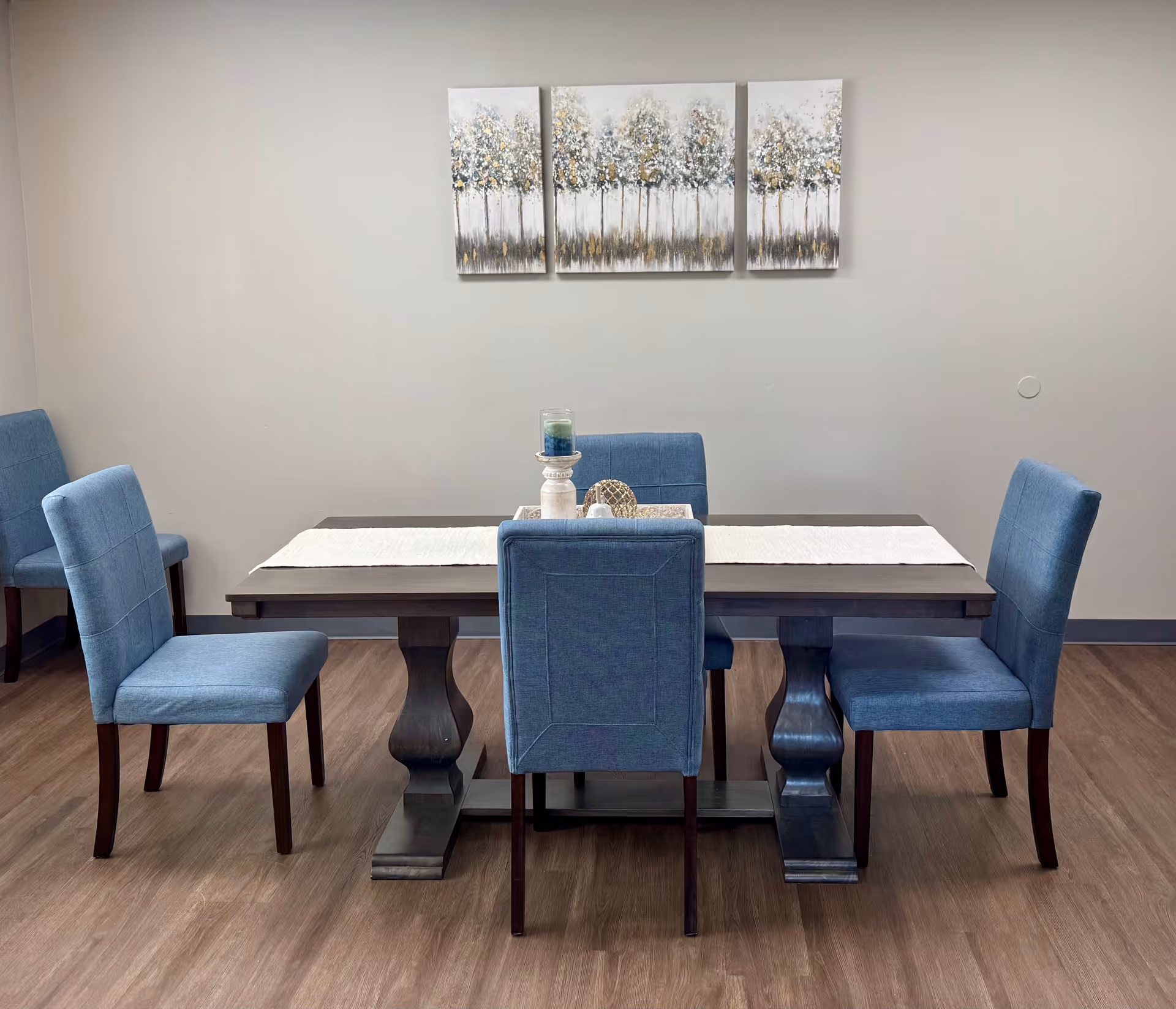 A dining area with a dark wooden rectangular table and six blue upholstered chairs. The table has a white runner and a decorative centerpiece with candles. On the wall above the table is a three-panel painting of abstract trees in shades of white, gray, and gold. The floor is wood, and the walls are light gray.