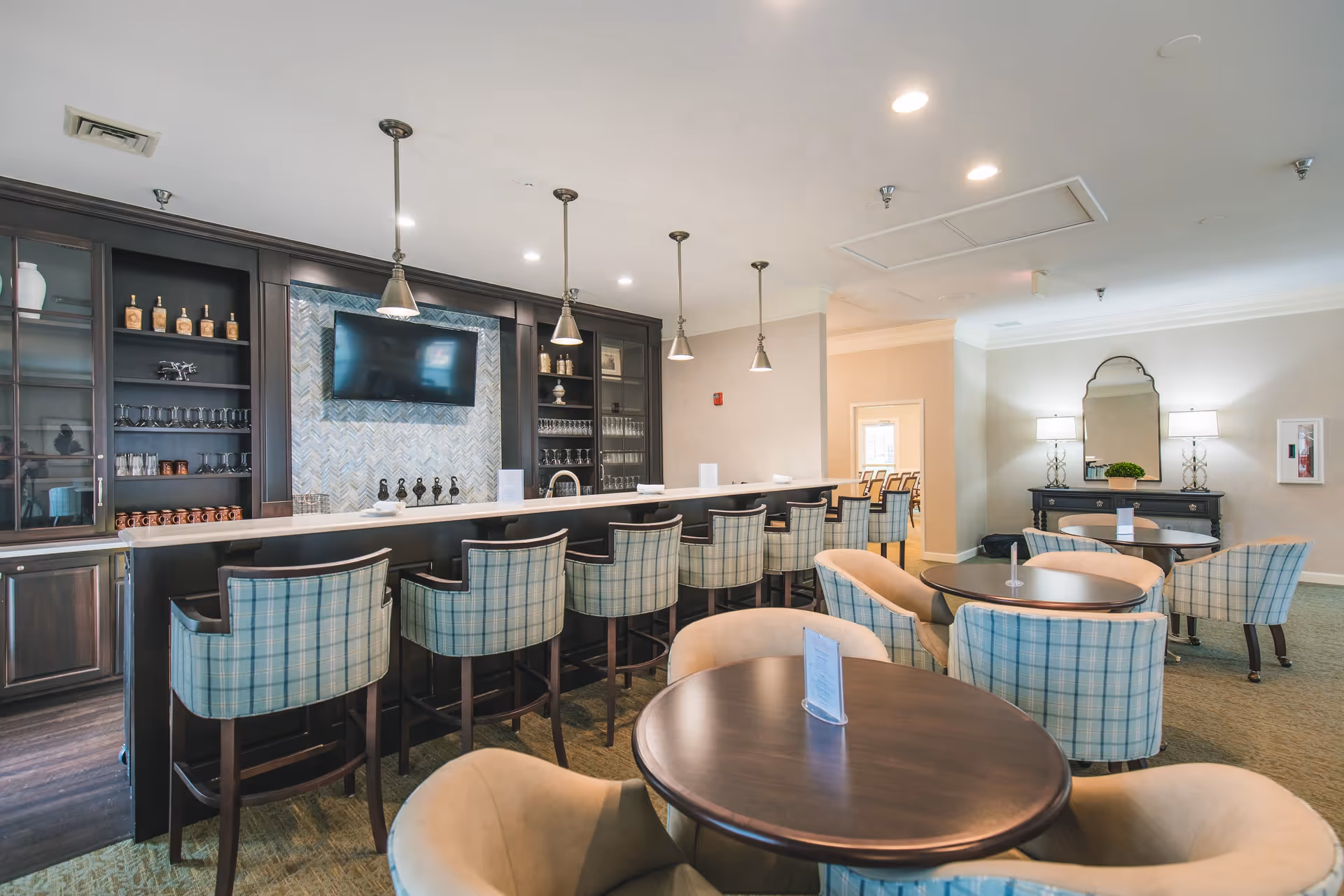 A spacious and well-lit common area with a long bar counter featuring several plaid upholstered bar stools. Behind the bar is a wall with shelves holding glassware and decorative items, and a mounted flat-screen TV. In the foreground, there are round tables surrounded by cushioned chairs, some with plaid upholstery. The room has a neutral color palette with beige walls, carpeted floors, and ceiling lights.
