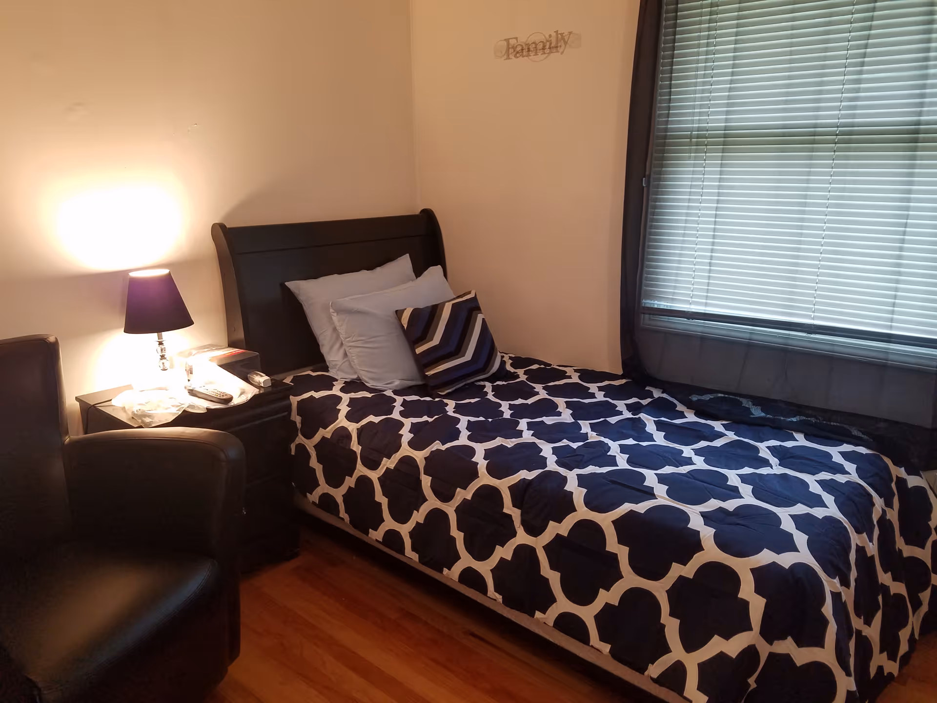 Small bedroom with a twin bed covered in navy-and-white patterned bedding, a nightstand with a lamp and chair beside a window.
