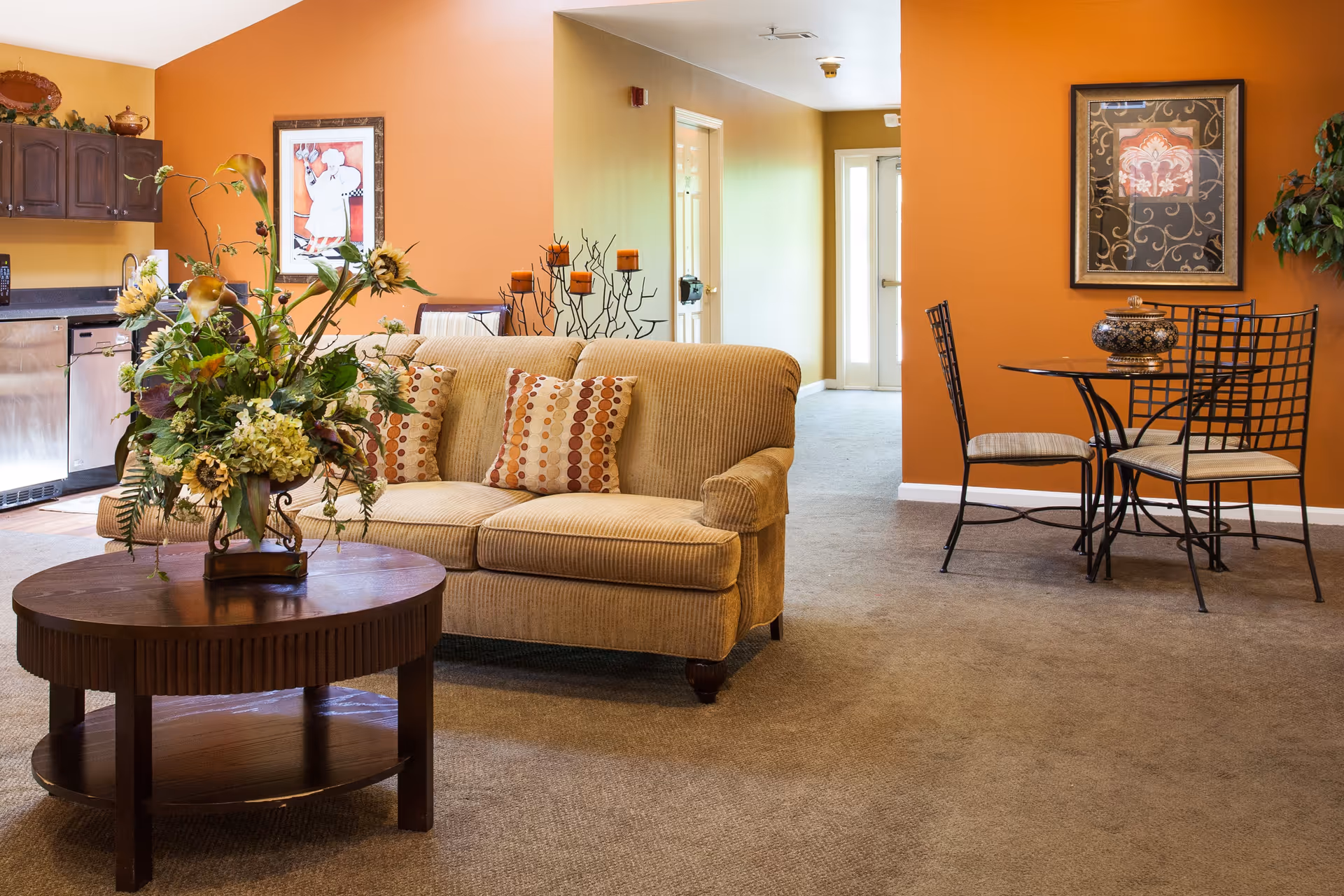 A cozy senior living common area with a beige sofa adorned with patterned cushions, a round wooden coffee table with a large floral arrangement, and a small dining table with four metal chairs. The walls are painted in warm orange and yellow tones, decorated with framed artwork and a candle holder. A kitchen area with dark wooden cabinets and stainless steel appliances is visible in the background.