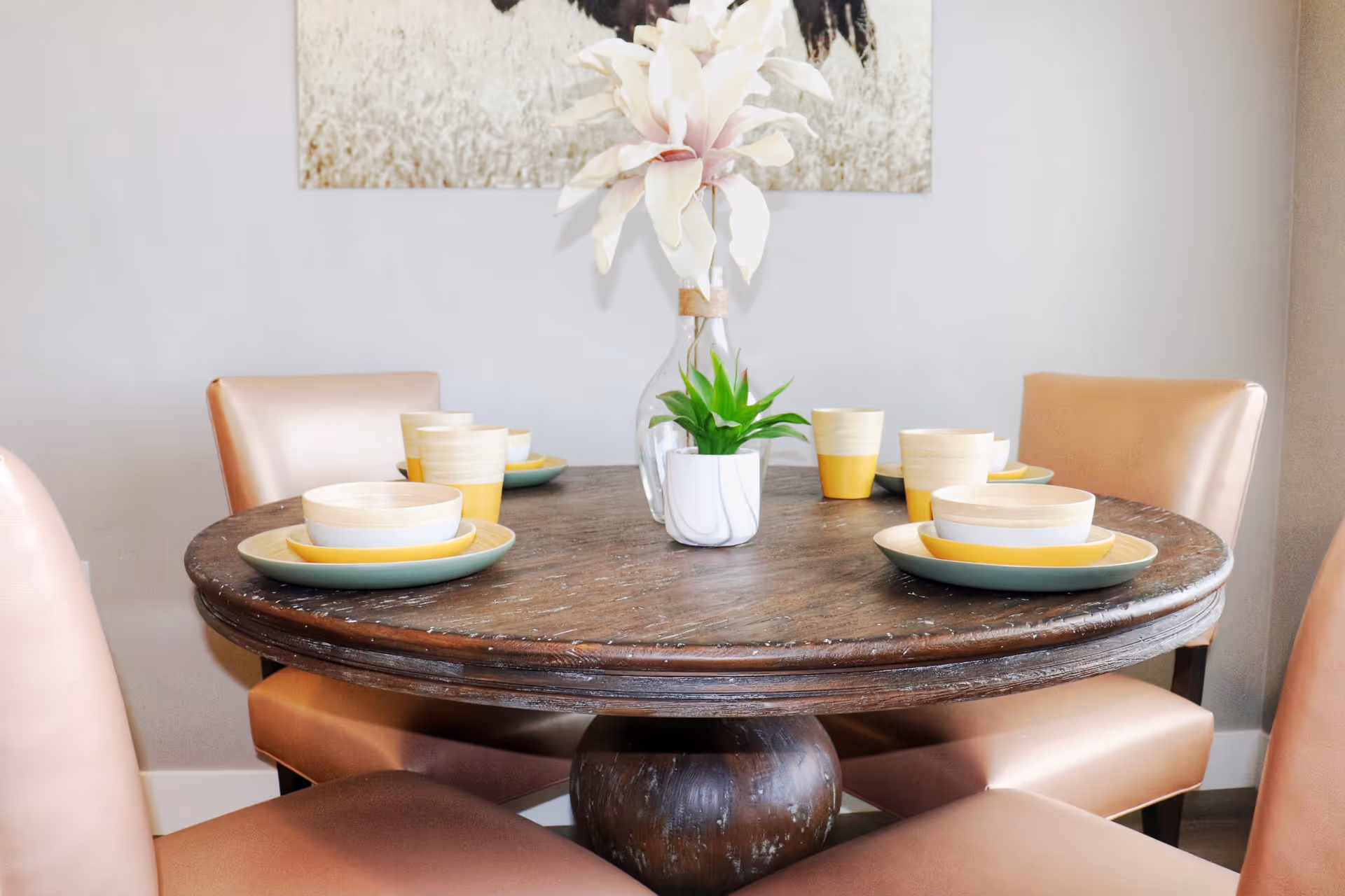 Round wooden dining table set with plates, bowls, cups and a floral centerpiece surrounded by upholstered chairs.