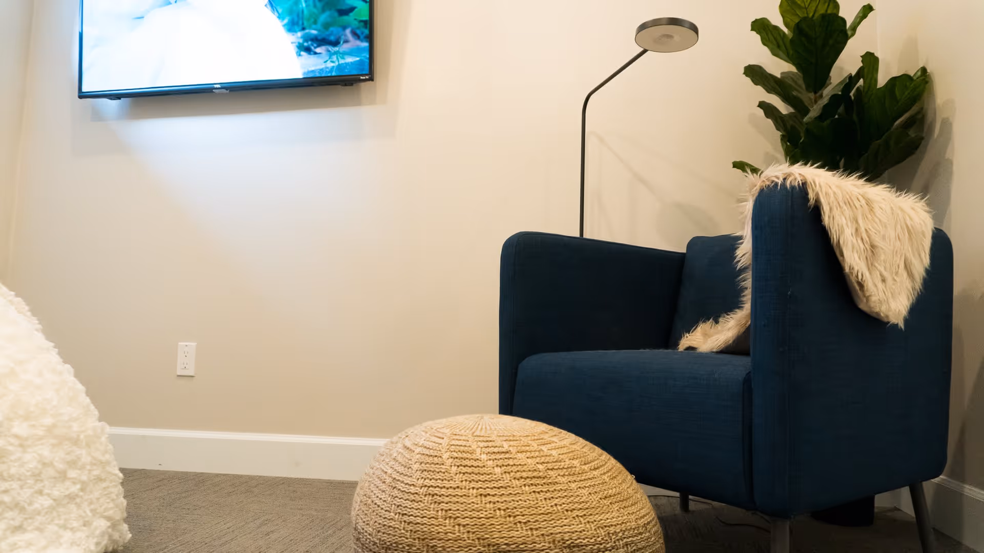 A cozy corner of a living room featuring a dark blue armchair with a light beige furry throw draped over its back. In front of the chair is a round, knitted beige pouf. A tall floor lamp with a circular shade stands behind the chair, and a large green leafy plant is placed in the corner. A flat-screen TV is mounted on the wall above the chair, and the room has light-colored walls and carpeted flooring.