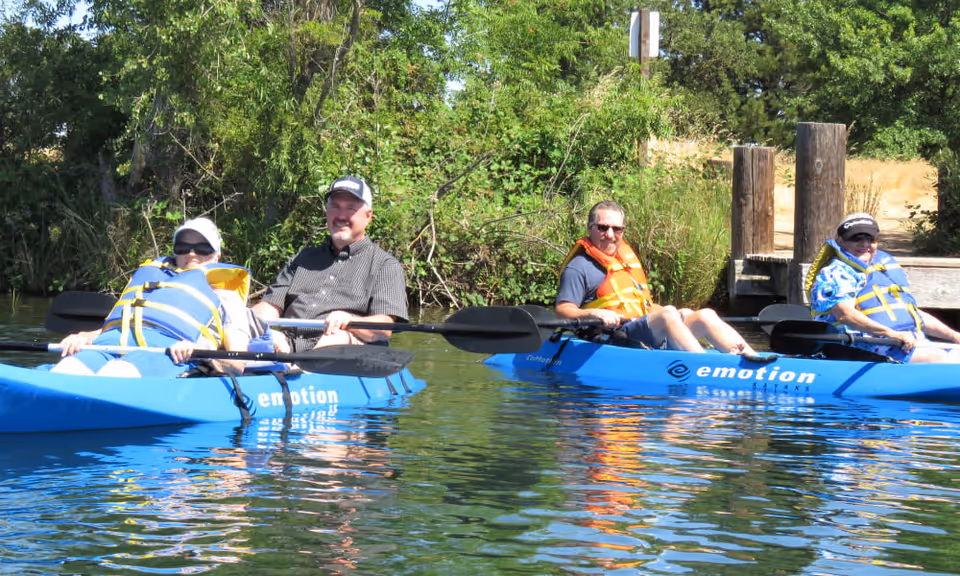Four adults wearing life jackets are kayaking on a calm body of water near a wooden dock with green trees and bushes in the background.
