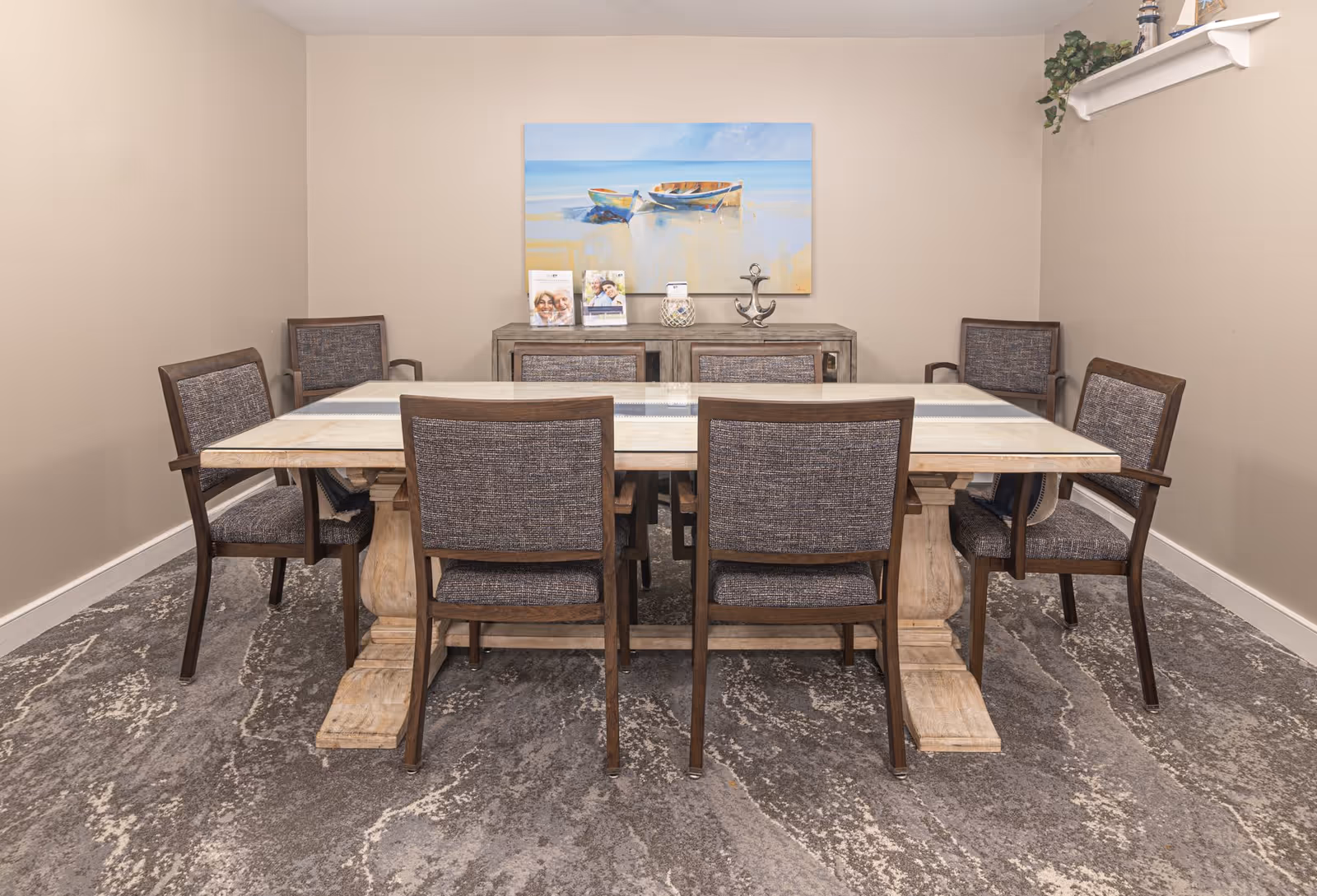 A dining room with a rectangular wooden table surrounded by six upholstered chairs. On the back wall, there is a sideboard with decorative items including brochures, a candle holder, and a metal anchor. Above the sideboard hangs a painting of two boats on a beach. The room has beige walls and a patterned carpeted floor.