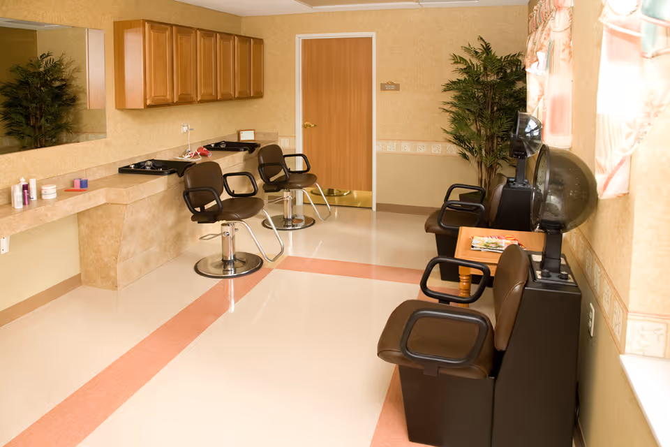 Interior view of a salon area in a senior living facility with two styling chairs in front of sinks and mirrors on the left side, and two brown chairs with hair dryers on the right side near a window with floral curtains. There is a wooden door at the back and a potted plant in the corner.