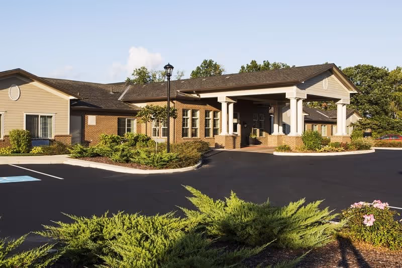 Front exterior of a single-story assisted living building with a covered porte-cochere entrance, landscaped beds, and a paved driveway.