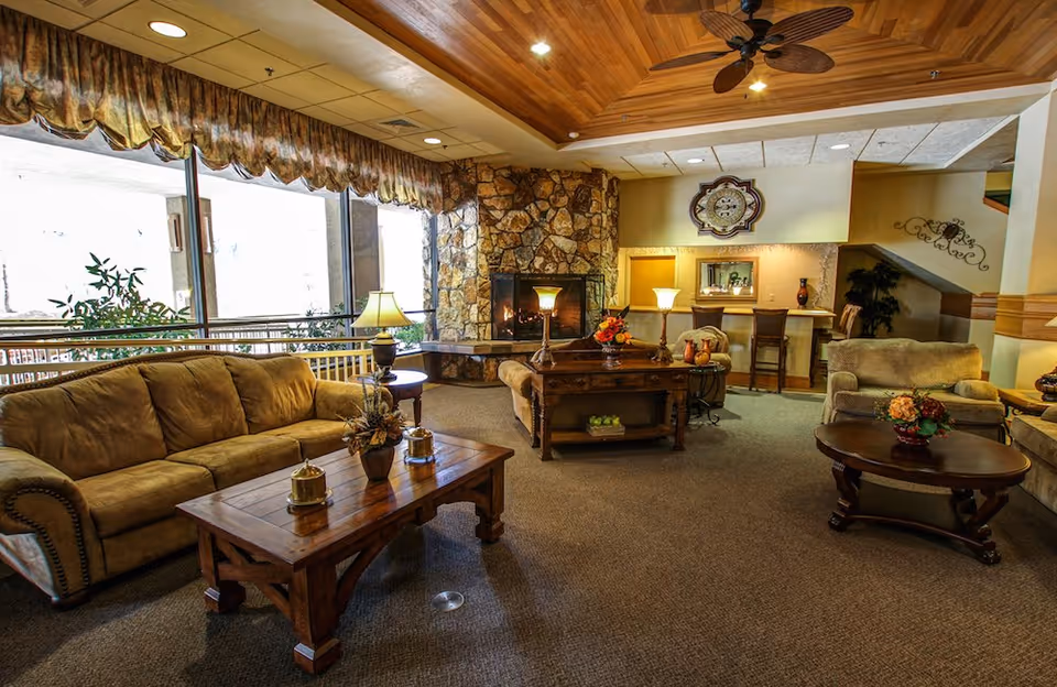 A cozy senior living facility common area with beige sofas and armchairs arranged around wooden coffee tables. There is a stone fireplace with a fire burning, lamps on tables, and a decorative clock on the wall. Large windows with patterned valances let in natural light, and the ceiling has wooden paneling with a ceiling fan.