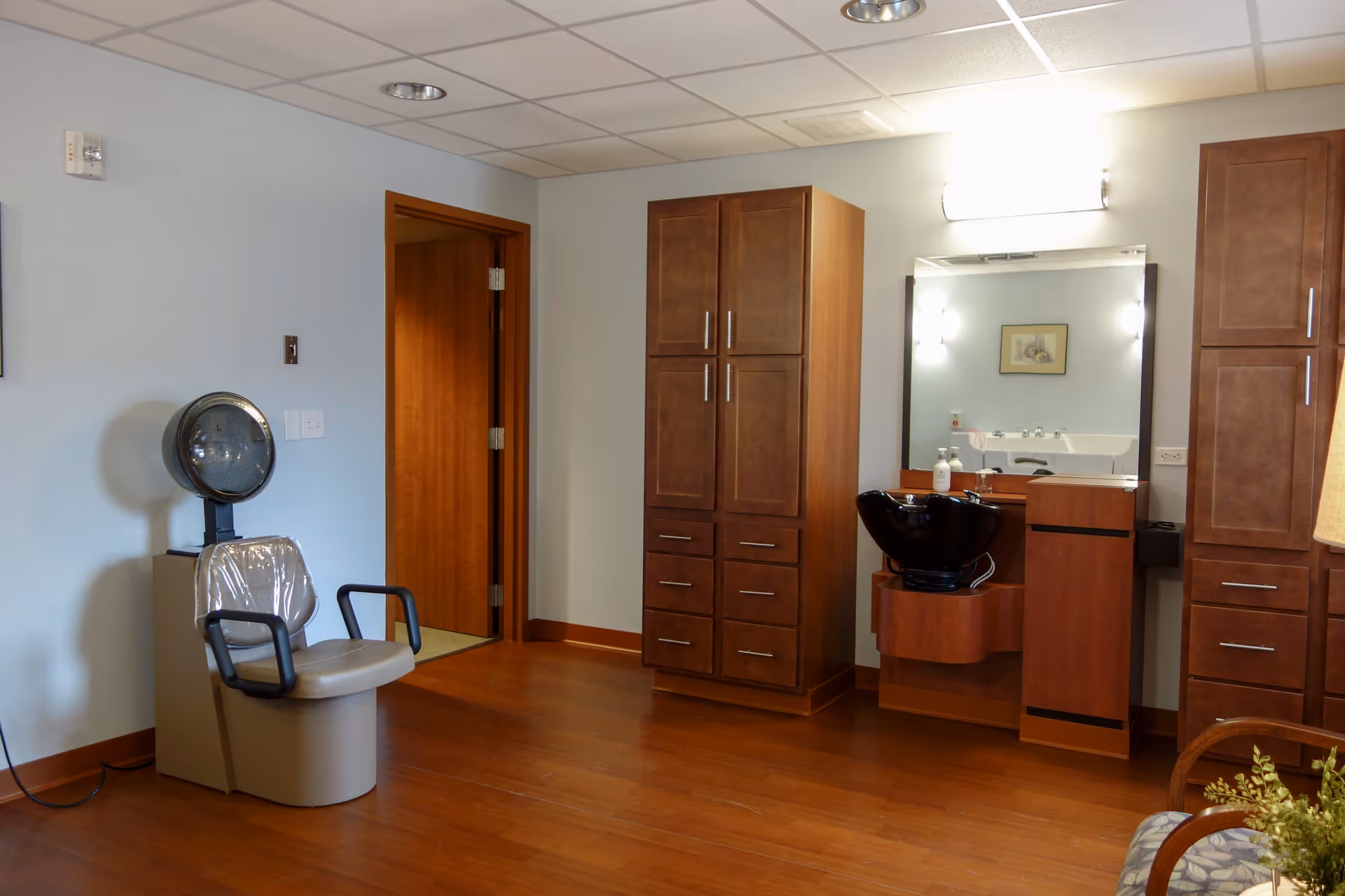 Interior view of a salon area in a senior living facility featuring a hair drying chair with a hood dryer, wooden cabinets, a black hair washing sink with a mirror above it, and a wooden floor.
