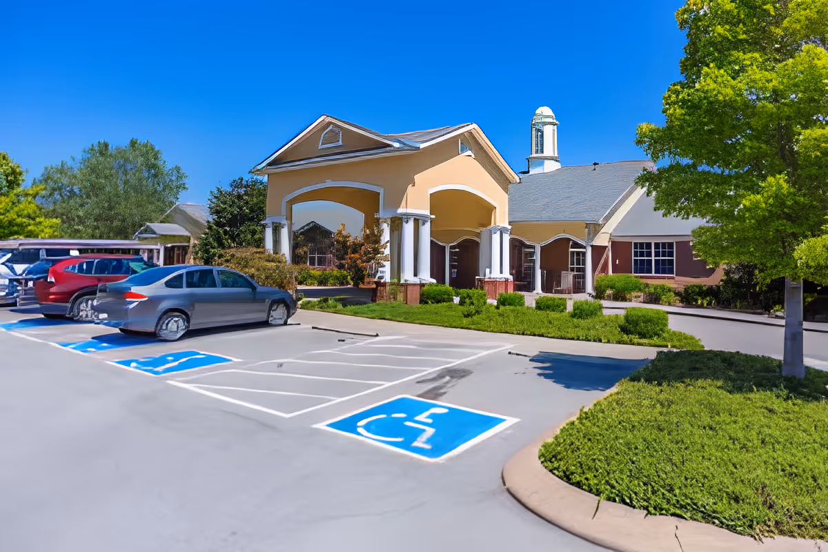 Exterior view of Charter Senior Living of Hermitage showing the entrance with a covered porch supported by white columns, a parking lot with several cars including handicapped parking spaces, and surrounding greenery under a clear blue sky.