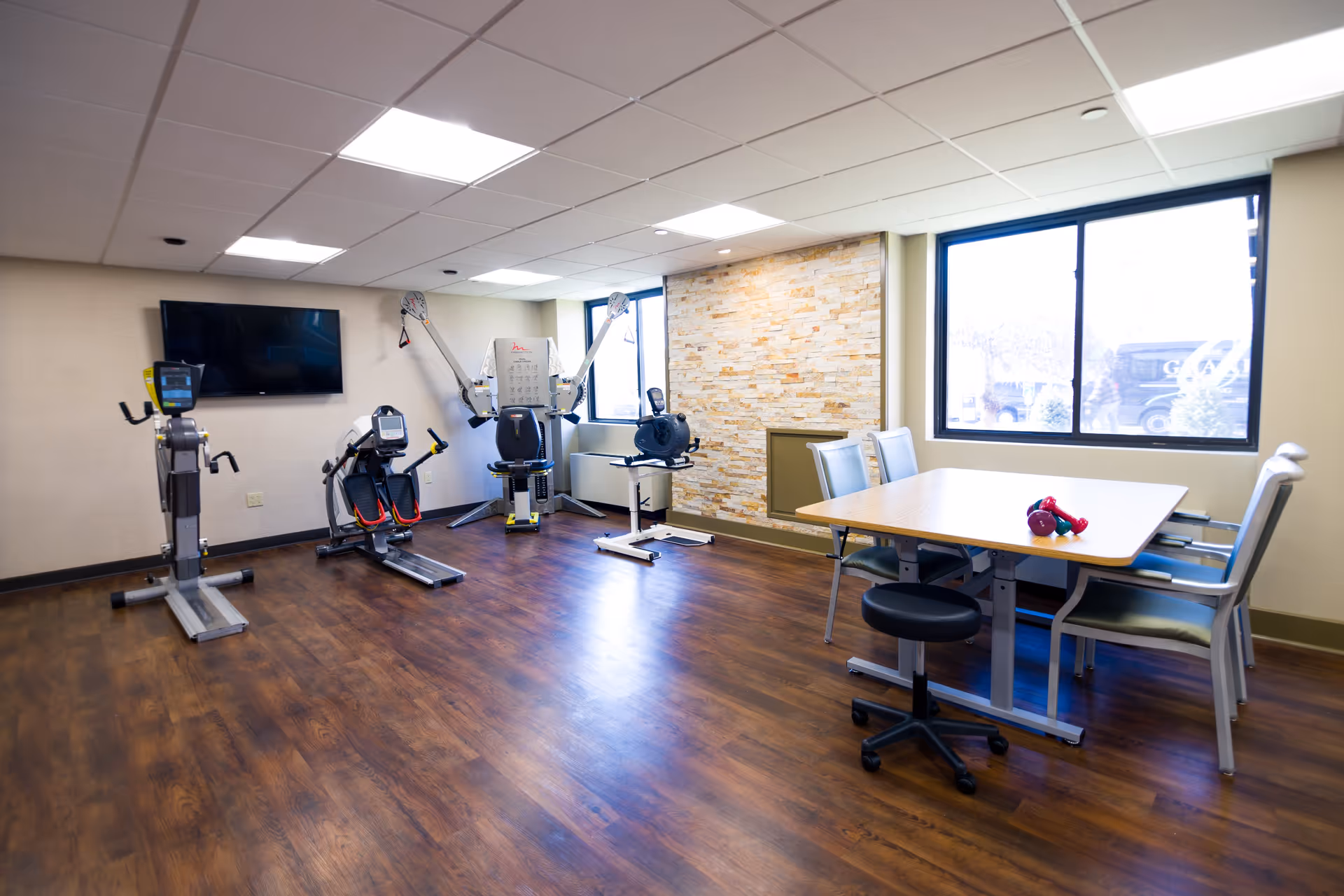 A rehabilitation room with exercise equipment including a rowing machine, stationary bike, and resistance machine. There is a wooden table with chairs and a stool near a large window letting in natural light. The room has wood flooring, a stone accent wall, and a mounted flat-screen TV.