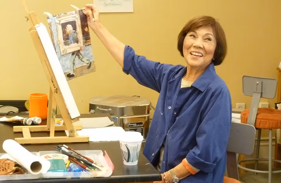 An elderly woman smiling while painting on a canvas set on an easel in an art room. The table in front of her holds various art supplies including paintbrushes, paper towels, and containers. The background shows a beige wall and some furniture.