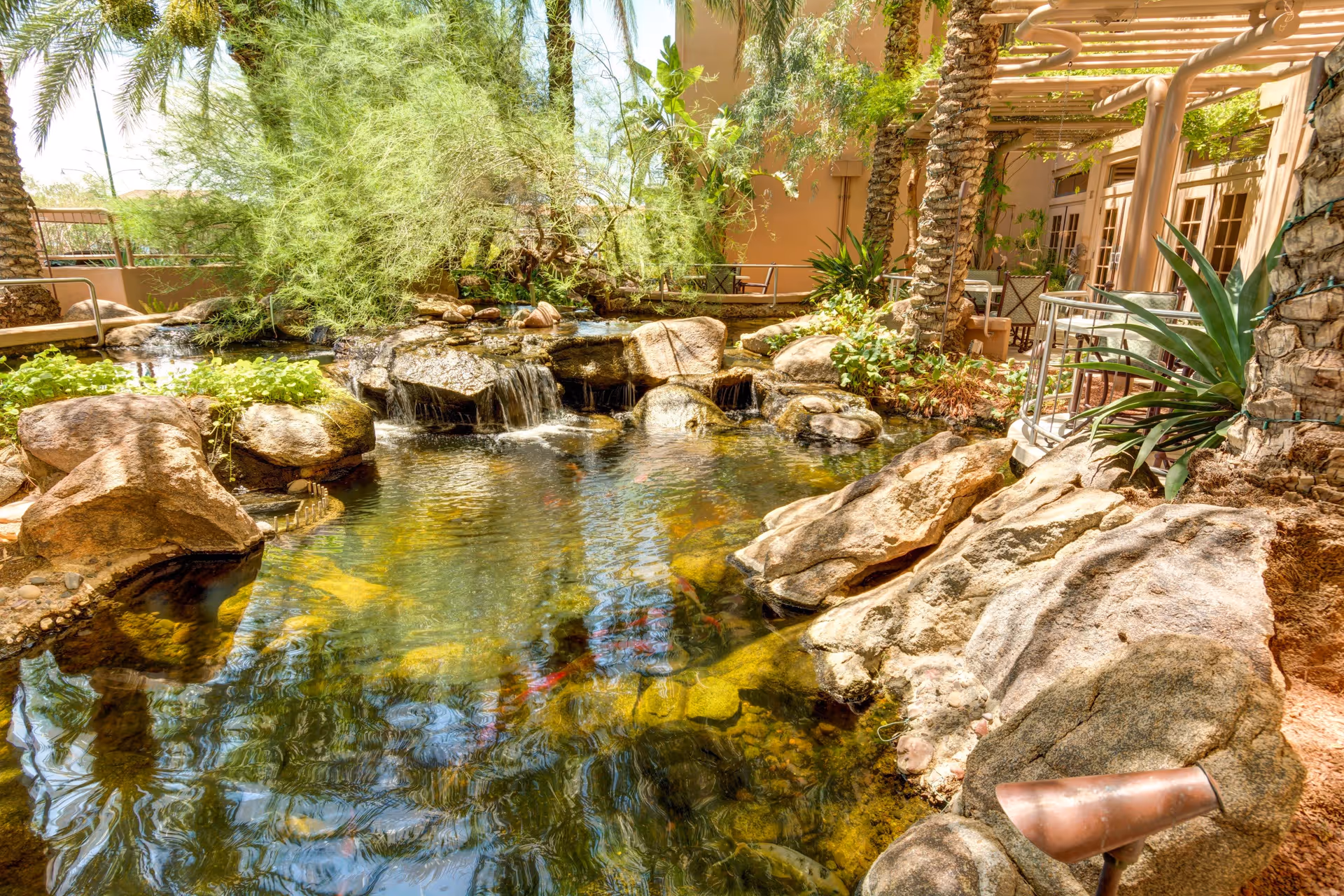 A serene outdoor garden area featuring a small pond with clear water, rocks, and a gentle waterfall surrounded by lush greenery and palm trees. There is a shaded seating area with tables and chairs under a pergola near a building.