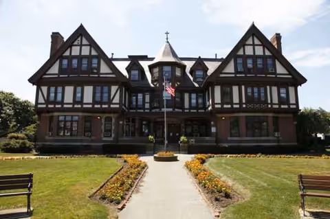 Tudor-style care facility building with central entrance, flagpole, and manicured gardens.