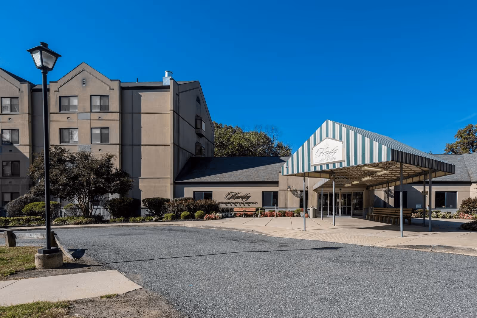 Exterior view of Kearsley Rehabilitation And Nursing Center building with a covered entrance, a lamp post, and surrounding landscaping under a clear blue sky.