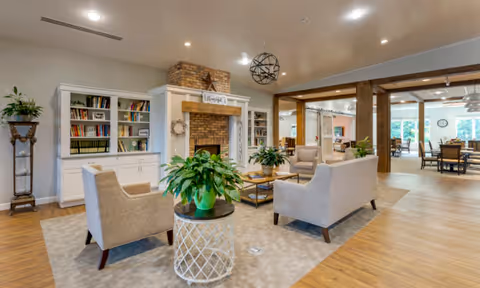 A spacious and well-lit living room area in a senior living facility featuring comfortable beige armchairs and sofas arranged around a coffee table with green plants. There is a brick fireplace with a wooden mantel and built-in white bookshelves on either side. The room has wooden flooring and a light-colored ceiling with recessed lighting. In the background, there is a dining area with tables and chairs near large windows letting in natural light.