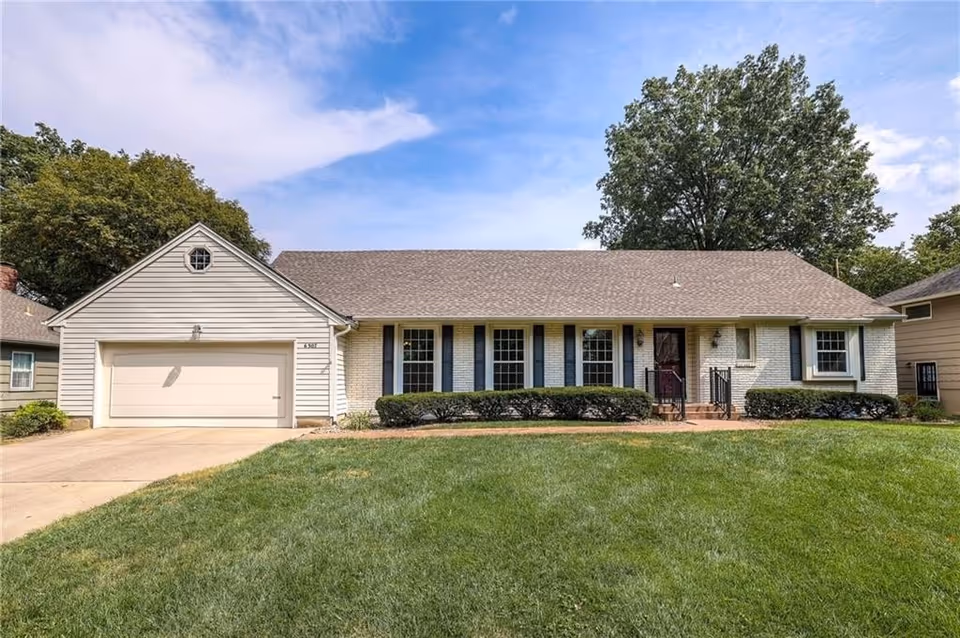 A single-story light-brick house with a two-car garage, a front walkway, and neatly trimmed shrubs along the entrance.