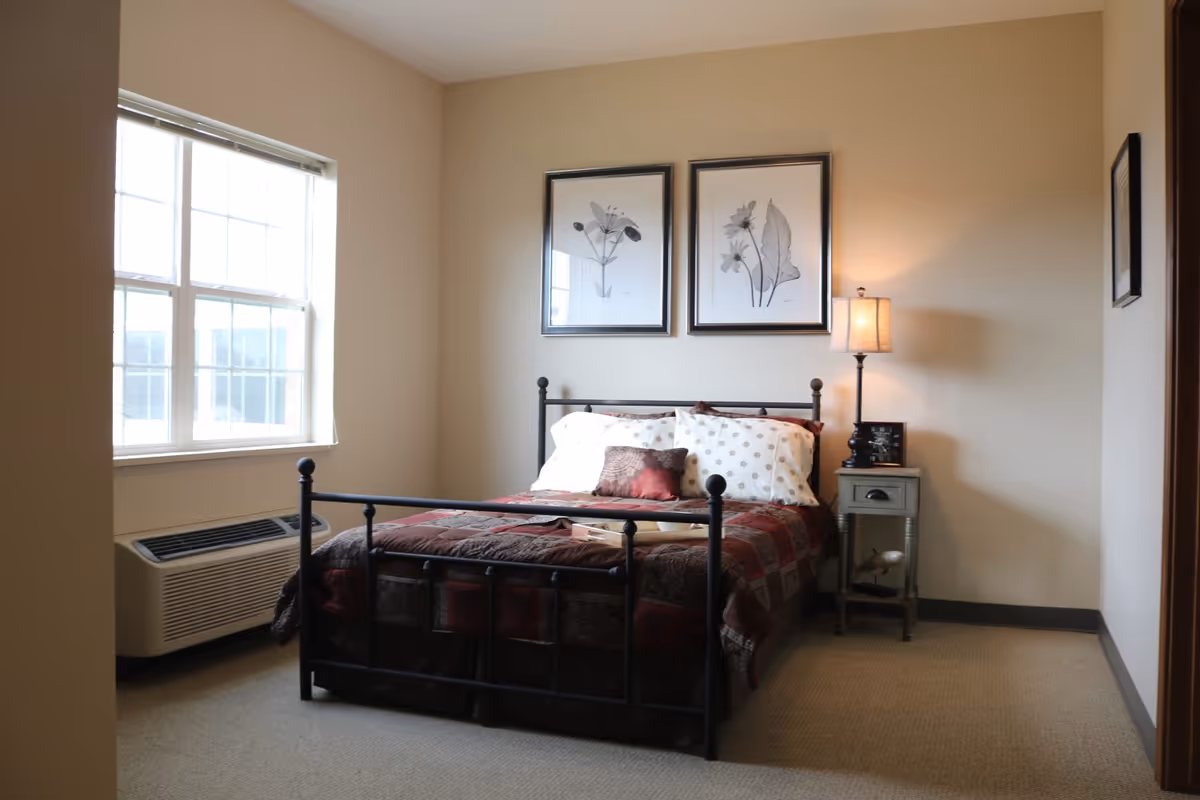 A cozy bedroom with a metal frame bed covered in a patterned quilt and pillows. There are two framed botanical prints hanging on the wall above the bed. A small nightstand with a lamp and a decorative item is placed to the right of the bed. A large window with blinds lets in natural light, and an air conditioning unit is installed below the window.