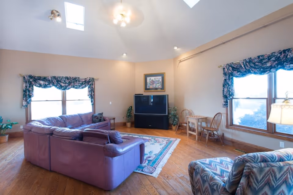A spacious living room with wooden floors, a large purple sectional sofa, a patterned armchair, and a floral area rug. The room has two large windows with floral valances, a small wooden table with two chairs, a large TV on a stand, and a framed floral painting on the wall. The ceiling is vaulted with skylights and recessed lighting.