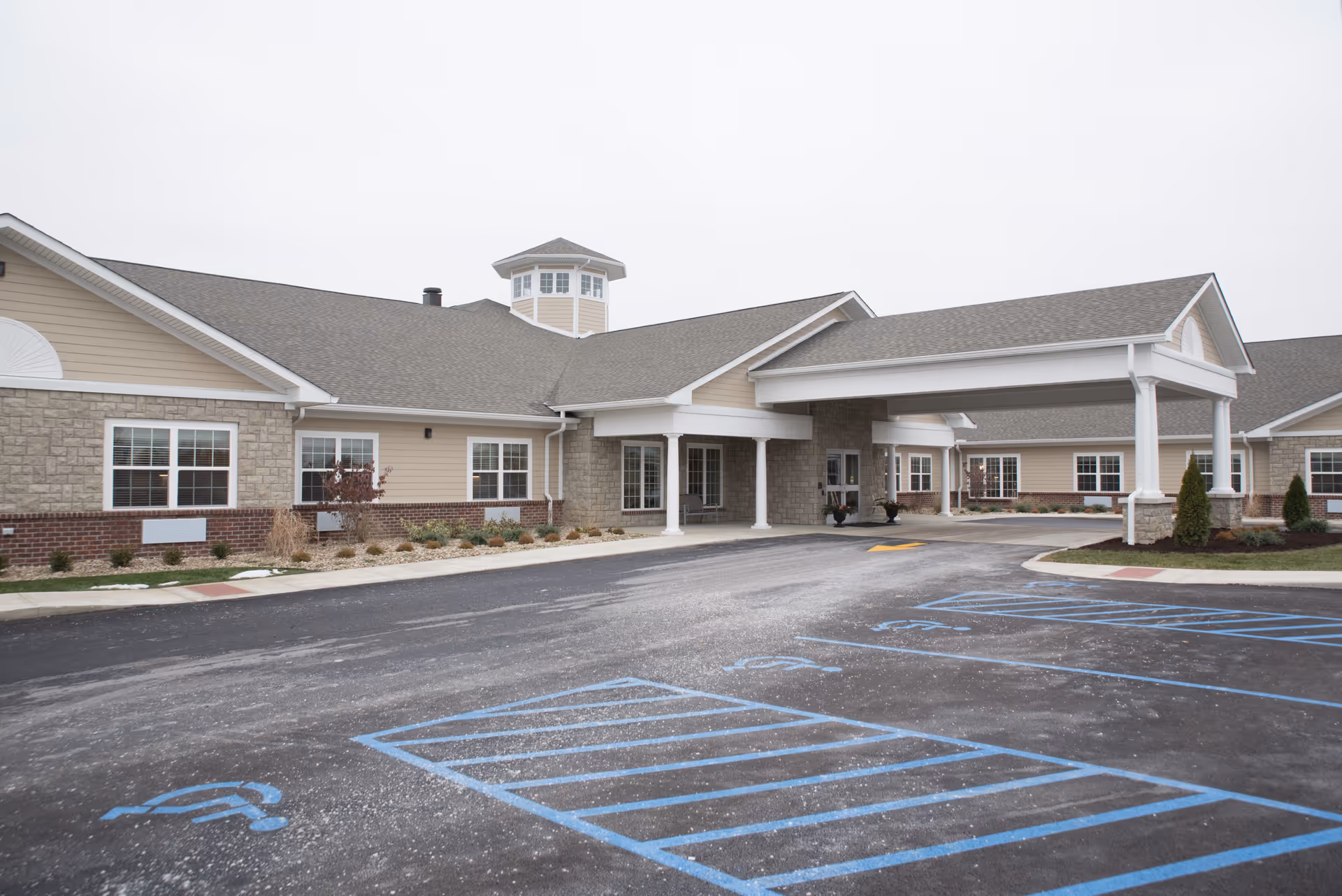 Exterior view of The Willows at Tiffin senior living facility showing a single-story building with beige siding, stone and brick accents, multiple windows, and a covered entrance with white columns. The foreground features a paved parking lot with blue handicap parking spaces.