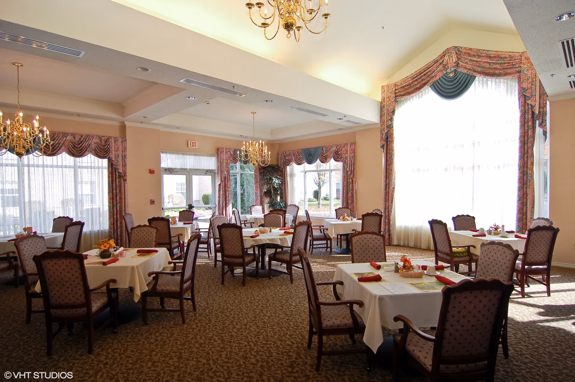 A bright dining room with several tables covered in white tablecloths, each set with red napkins and small floral centerpieces. The room features large windows with patterned drapes, chandeliers hanging from the ceiling, and upholstered chairs around the tables.