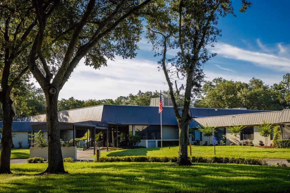 Exterior view of Solaris HealthCare Plant City building with a blue roof, surrounded by green grass and trees under a partly cloudy sky. An American flag is visible near the entrance.