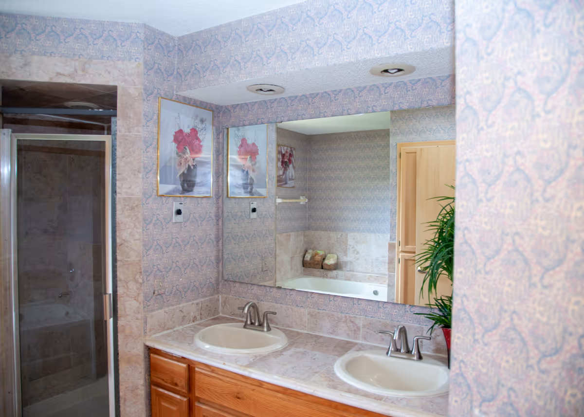 Double-sink bathroom vanity with a large mirror, shower stall, bathtub and patterned wallpaper.