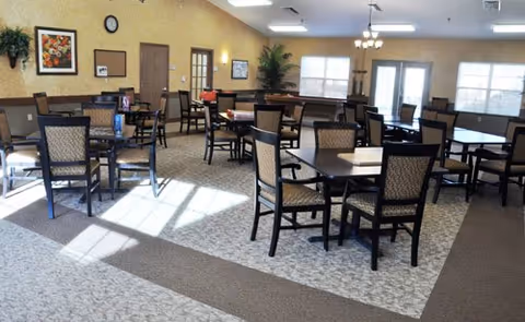 A spacious dining room with multiple square tables and cushioned chairs arranged neatly. The room has beige walls adorned with framed artwork and a clock. Large windows and glass doors allow natural light to brighten the space. There are ceiling lights and a carpeted floor with a patterned design.